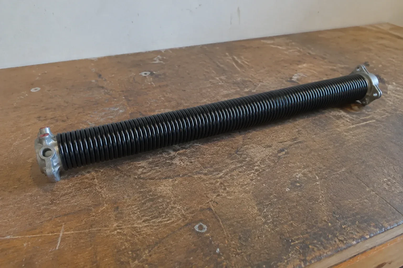 Detached black coil spring with metal end fittings on a workbench. — Torsion Spring Replacement, Grimsby